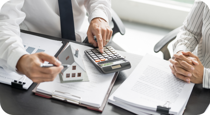 Two people sitting at a desk with property documents, a calculator, and a small model house, discussing financial details.
